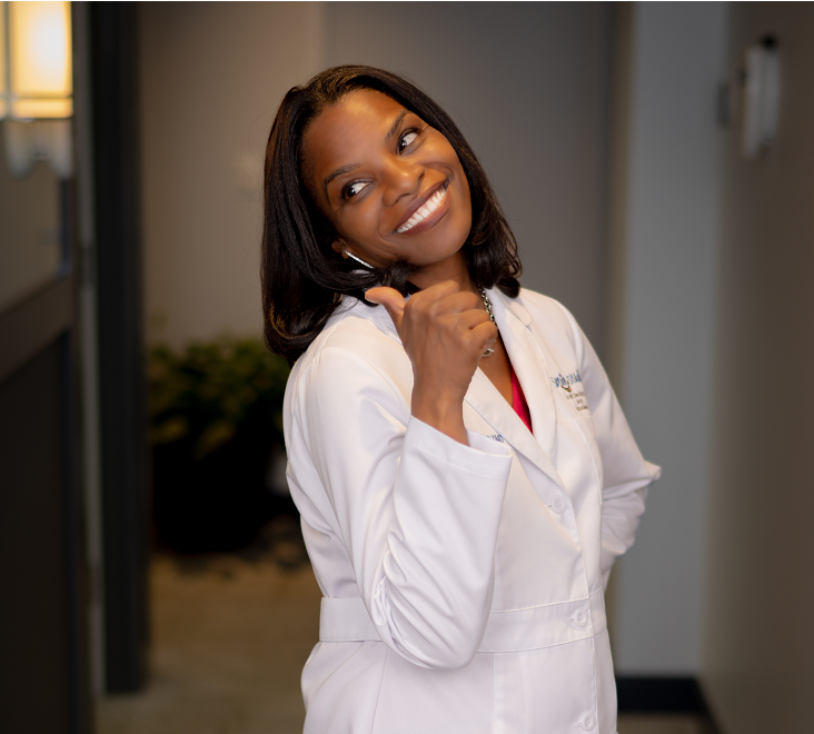 Smiling woman in a white lab coat stands in a hallway, exuding confidence and warmth. Her gesture is welcoming, with soft lighting creating an inviting atmosphere.