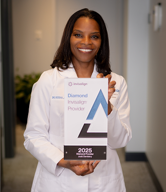 A smiling woman in a lab coat holds a "Diamond Invisalign Provider 2025" award. The setting is a professional office hallway, conveying success and professionalism.