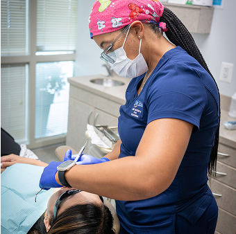Dentist wearing a pink cap and blue scrubs works on a patient in a dental chair. The room is bright with a calm, focused atmosphere.
