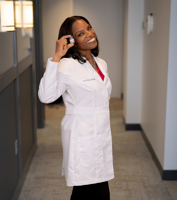 A smiling woman in a white lab coat holds a small bottle, suggesting a healthcare or scientific setting. The hallway is softly lit, conveying a professional tone.