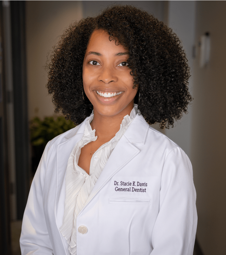 Smiling woman with curly hair in a white dentist coat stands in a professional setting, exuding confidence and friendliness.